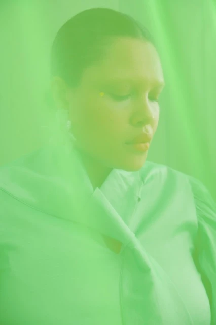 Close-up portrait of a woman with closed eyes, luminous skin, and bright yellow makeup in a monochromatic green environment with soft lighting.