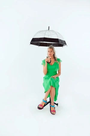 Adult woman in a green dress sits on a stool indoors holding a clear umbrella against a white background in a studio shot.