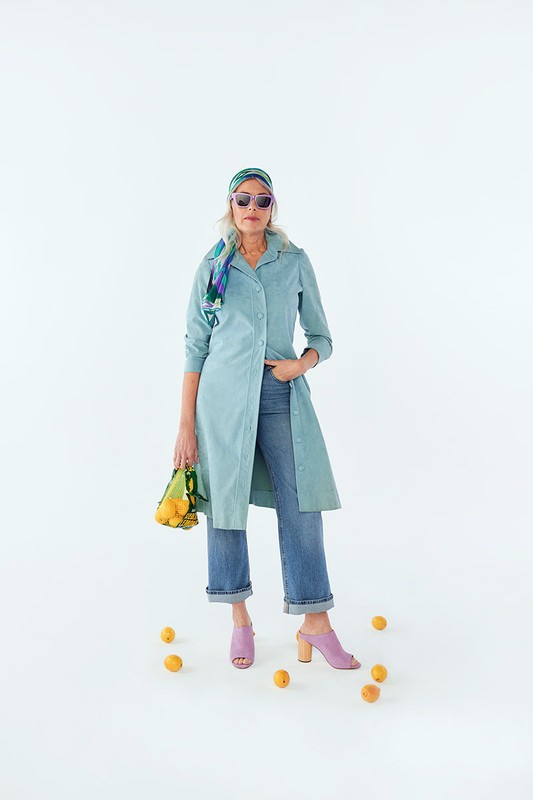 A woman in blue dress, jeans, and mules poses in a photo studio surrounded by lemons, holding a net bag also filled with lemons.