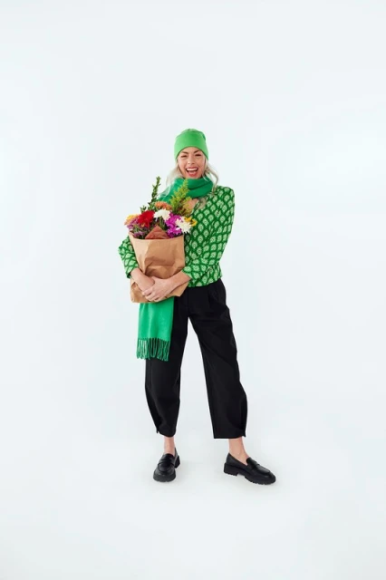 A happy mature woman in a green patterned sweater, scarf, and beanie holds a brown paper bag with flowers, standing against a white backdrop.