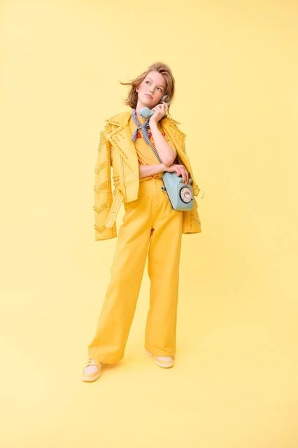 Full-length shot of a young woman in yellow outfit, holding a blue retro telephone, looking up, in a yellow studio environment.