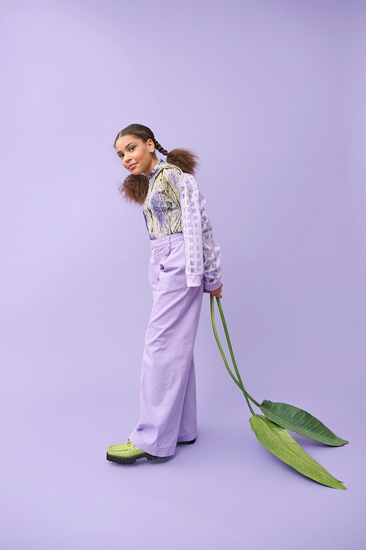 Young female fashion model in lilac pants and patterned top, holding green leaves, posing in a studio with a lilac background.