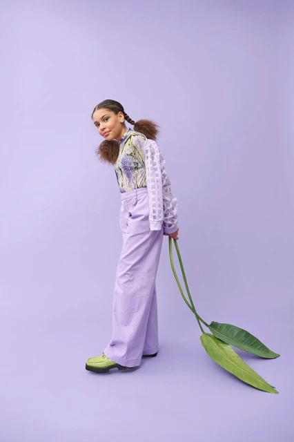 Young female fashion model in lilac pants and patterned top, holding green leaves, posing in a studio with a lilac background.
