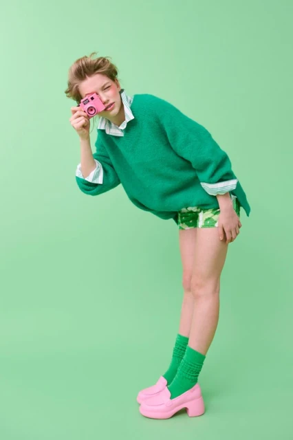 Young adult woman in green sweater poses with a vintage-style pink camera, standing against a green background in a studio environment.