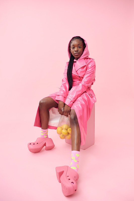 Young woman in a pink hooded coat and platform shoes sits on a pink cube holding lemons against a matching pink backdrop in a studio.
