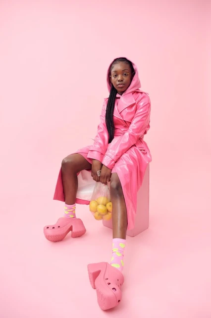 Young woman in a pink hooded coat and platform shoes sits on a pink cube holding lemons against a matching pink backdrop in a studio.