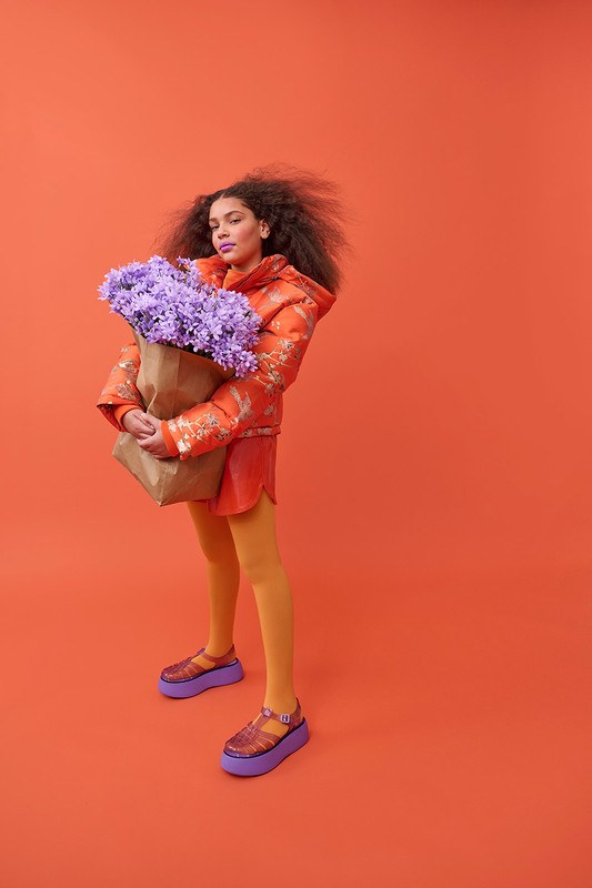 Woman in an orange jacket and yellow tights holding a brown paper bag with purple flowers against a matching orange backdrop.