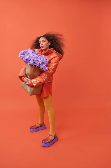 Woman in an orange jacket and yellow tights holding a brown paper bag with purple flowers against a matching orange backdrop.