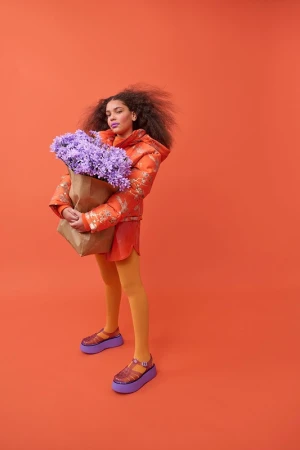 Woman in an orange jacket and yellow tights holding a brown paper bag with purple flowers against a matching orange backdrop.