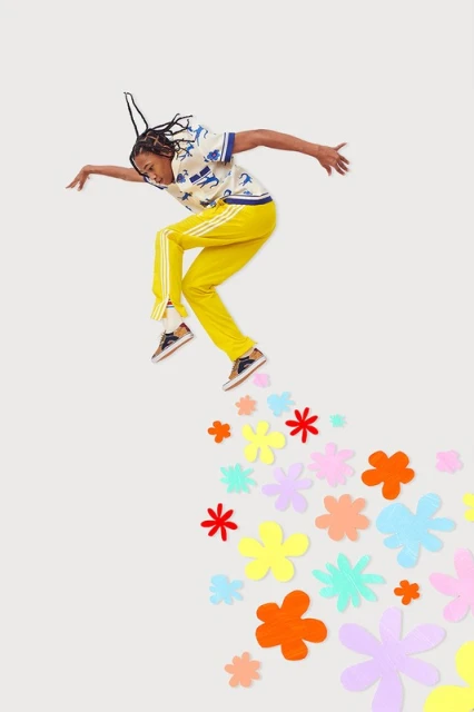 Full shot of a young adult male dancer with yellow pants jumping on a white background with colorful paper flowers. Fashion, movement.