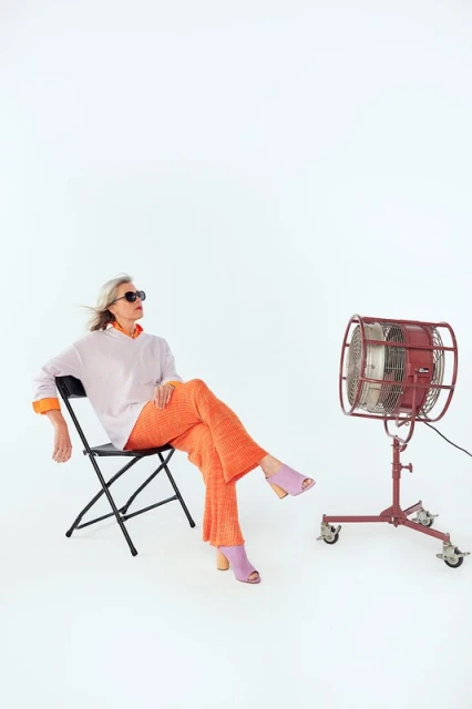 Adult woman with grey hair in sunglasses sitting on a chair next to a vintage fan in a bright photo studio with white background.