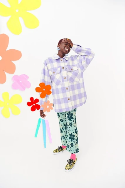A black woman is smiling, wearing a patterned flannel and pants, and holding paper flowers in a high-key, well-lit studio shot.