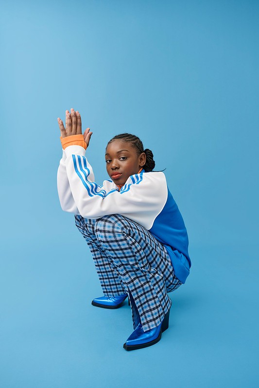 A young woman with braided hair poses in a squatting position against a light blue seamless backdrop, wearing blue plaid pants and shiny blue boots.