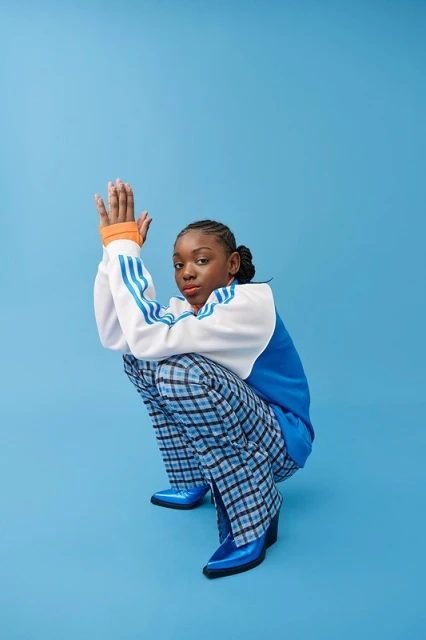A young woman with braided hair poses in a squatting position against a light blue seamless backdrop, wearing blue plaid pants and shiny blue boots.
