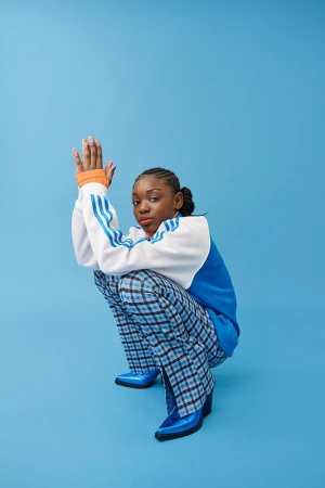 A young woman with braided hair poses in a squatting position against a light blue seamless backdrop, wearing blue plaid pants and shiny blue boots.