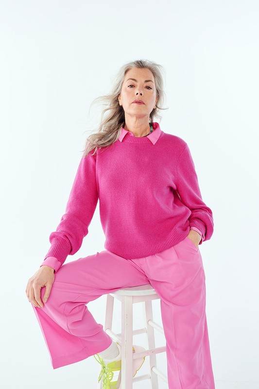 Elegant older woman in bright pink sweater and wide-leg pants sits on a white stool in a high-key fashion studio shot.