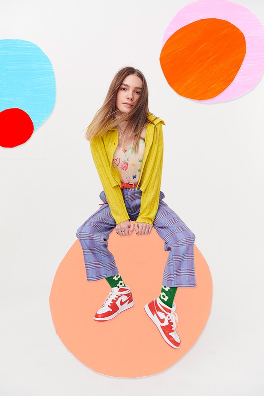 A young woman in a colorful outfit sits on a large orange circle, with painted abstract circles in the background, in a studio setting.