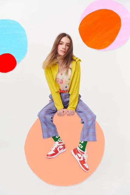 A young woman in a colorful outfit sits on a large orange circle, with painted abstract circles in the background, in a studio setting.