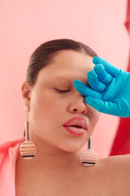 Woman with closed eyes wearing large earrings and a blue glove, posing in front of a pink backdrop and a red cloth.
