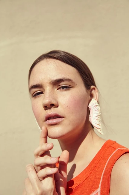 Close-up portrait of a young adult female wearing large fringed earrings, an orange top, and a neutral backdrop in soft, diffused light.