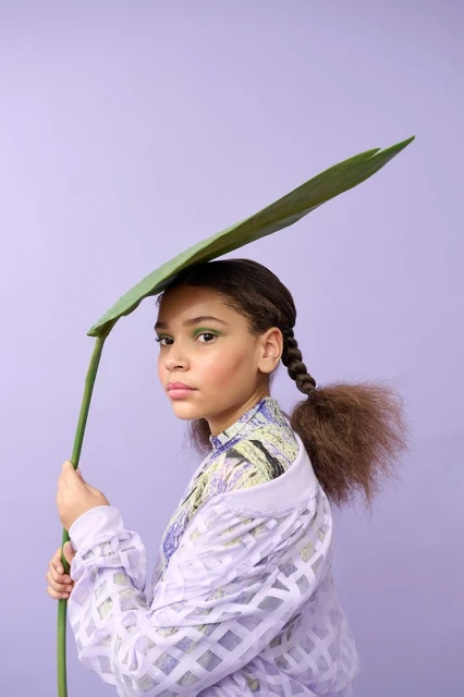 A child poses in a lilac studio with a large leaf, wearing a patterned top and open jacket with braided hair and green eye shadow.