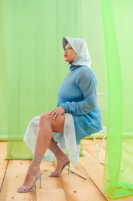 A woman in a blue dress and head covering sits on a chair against a green background in a studio fashion portrait with a side angle.
