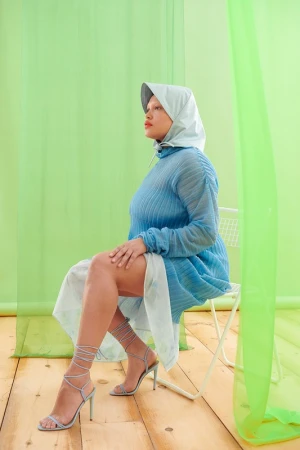 A woman in a blue dress and head covering sits on a chair against a green background in a studio fashion portrait with a side angle.