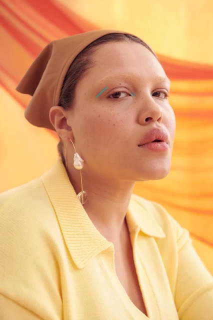 Portrait of a woman with a headscarf and modern jewelry, posing in front of an orange backdrop with abstract wave patterns.