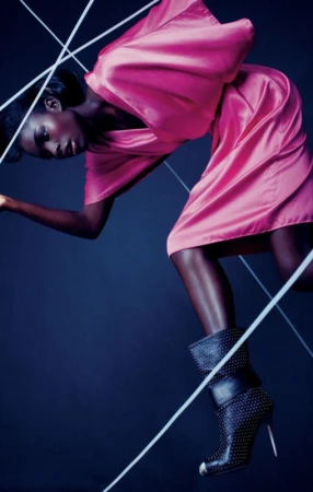 A Black female model in a pink satin dress and studded boots poses against a dark background amidst white string, high fashion.
