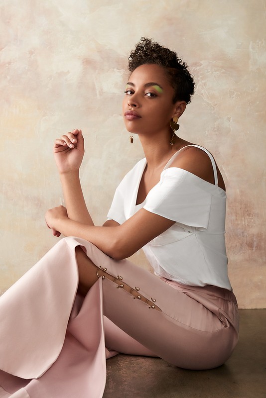 Elegant woman in white top and pink pants sits in a studio with soft lighting. Eye-level, medium shot with a textured background.