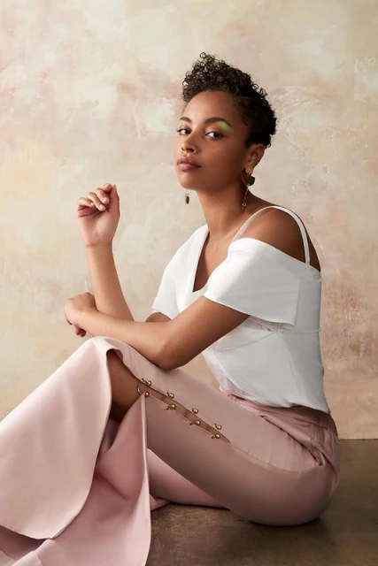 Elegant woman in white top and pink pants sits in a studio with soft lighting. Eye-level, medium shot with a textured background.