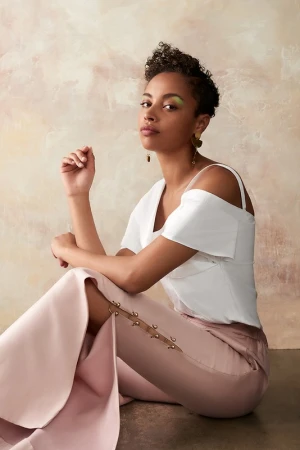 Elegant woman in white top and pink pants sits in a studio with soft lighting. Eye-level, medium shot with a textured background.