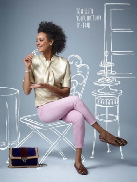 A smiling woman with curly hair sits on a chair next to a handbag in front of a gray background with white line art drawings.