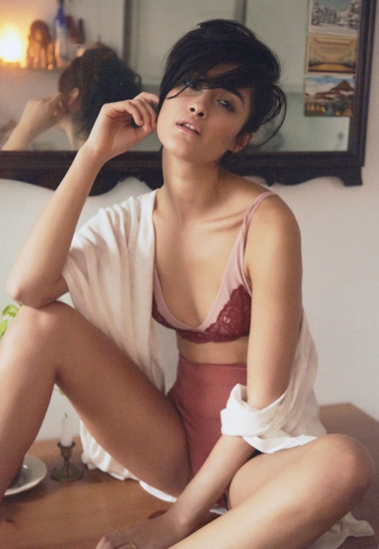 Portrait of a young woman sitting cross-legged on a table, wearing lingerie and a shirt, with a mirror reflecting a room in the background.