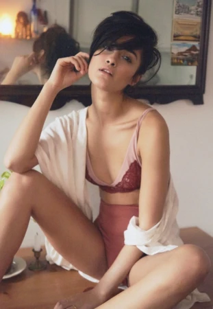Portrait of a young woman sitting cross-legged on a table, wearing lingerie and a shirt, with a mirror reflecting a room in the background.