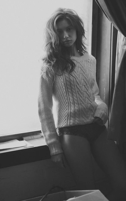 Black and white shot of a woman in a sweater sitting near a window with natural light, exuding a natural and slightly somber mood.
