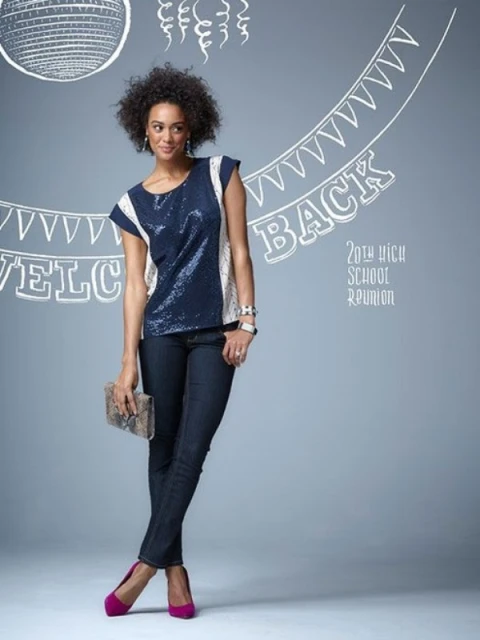 Full-length shot of a woman in a blue sequin top and dark jeans, holding a clutch, posing against a light gray background.