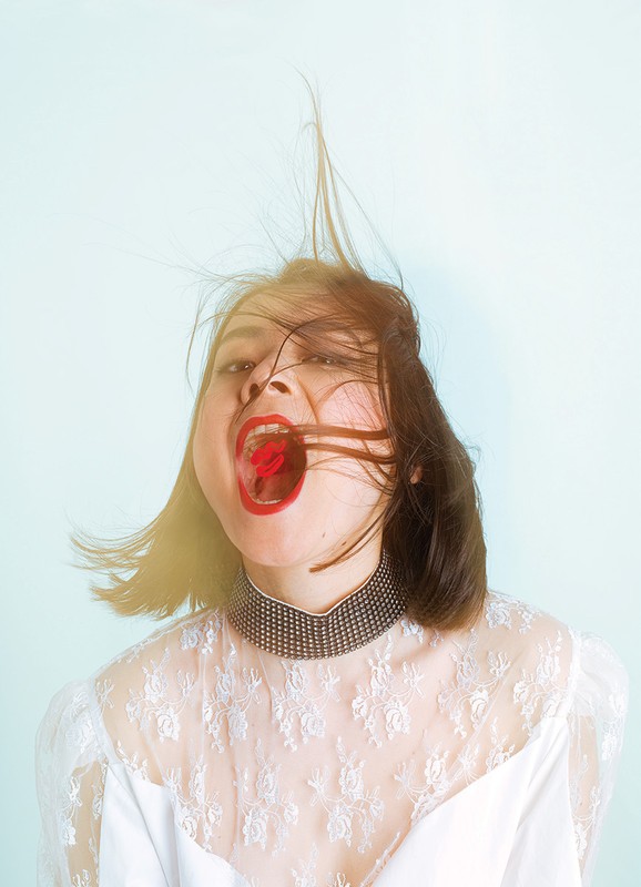 Portrait of a woman with red lipstick holding a red cherry in her mouth, wearing a white lace dress against a light blue background.
