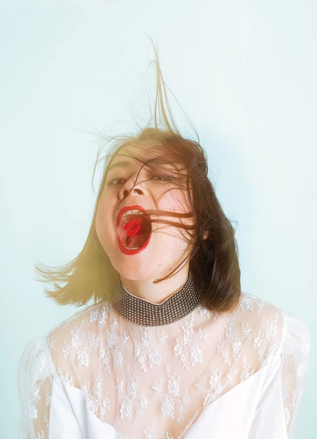 Portrait of a woman with red lipstick holding a red cherry in her mouth, wearing a white lace dress against a light blue background.