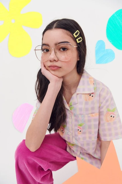 Young girl with glasses and hair clips poses in a patterned blouse with colorful illustrated background elements in a studio setting.