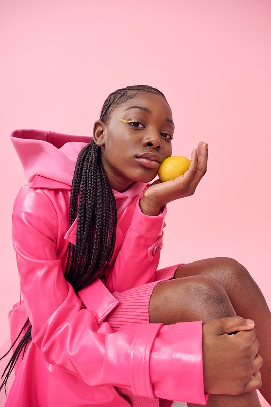 Portrait of a young Black woman with yellow eyeliner and braids, holding a lemon against a pink background, wearing a pink jacket.
