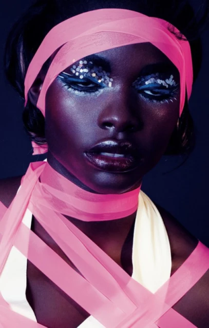 Close-up of a woman with dark skin and creative makeup, featuring glitter eyeshadow, glossy lips, and a pink ribbon head wrap and body wrap.