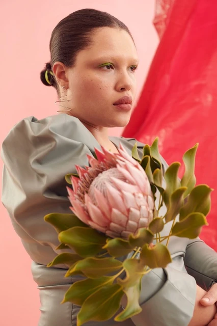 Portrait of a woman in a grey dress holding a protea flower against a pink backdrop with red fabric draping in the background.