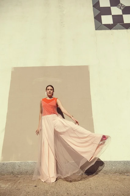 A woman in an orange top and flowing peach skirt poses in front of a beige wall with geometric black and white tiles overhead.