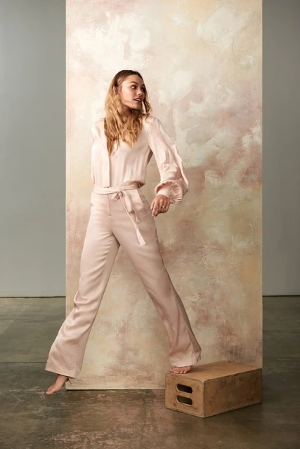 Woman poses in pink satin top and pants, standing on a wooden box in front of a painted canvas in a photo studio.