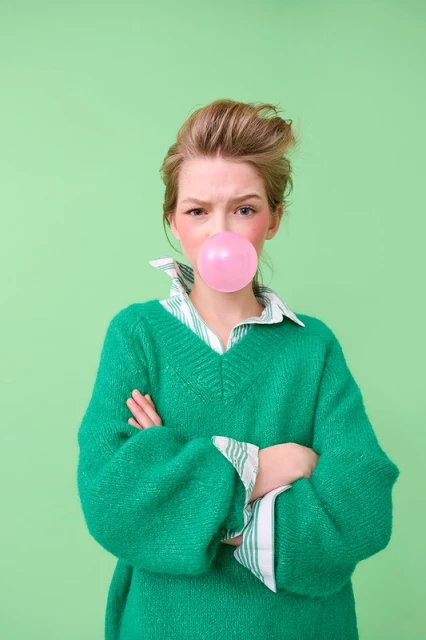 A young woman in a green sweater blows a pink bubble with gum against a light green backdrop, exuding a youthful and playful vibe.