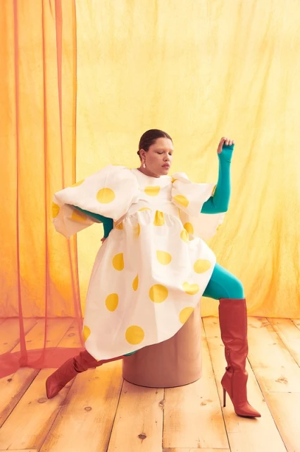 A woman with fair skin poses in a yellow-spotted white dress, turquoise tights, and red boots against a yellow-orange backdrop.