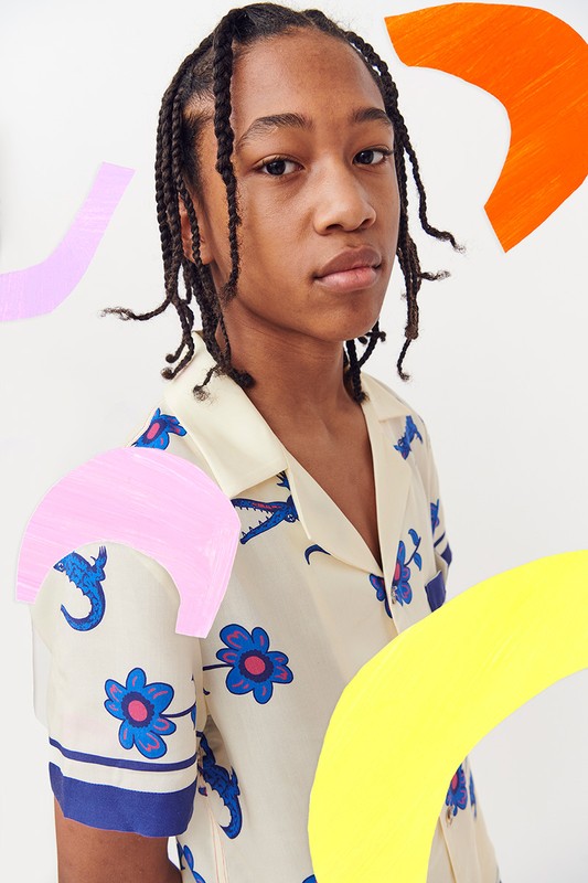 Portrait of a young person with braided hair wearing a patterned collared shirt, framed by abstract shapes in yellow, pink, and orange.