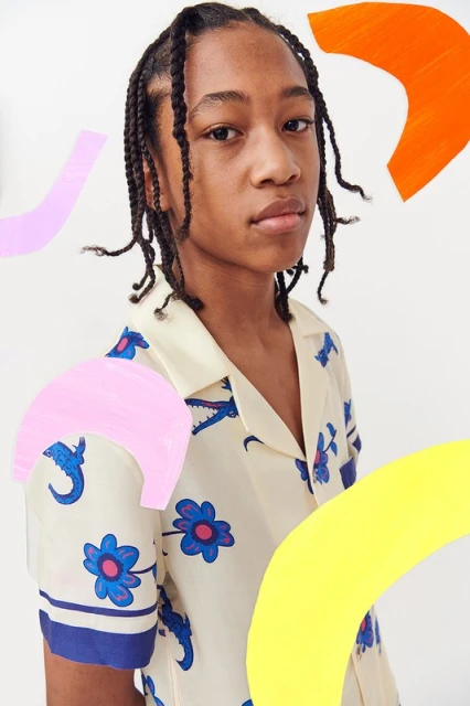Portrait of a young person with braided hair wearing a patterned collared shirt, framed by abstract shapes in yellow, pink, and orange.