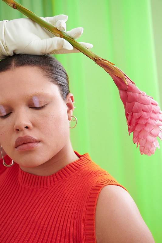 Portrait of an adult female with closed eyes, wearing an orange sleeveless top, posed with a pink flower and white gloves against green.